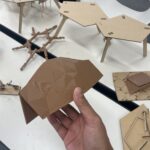 Close-up of a 3D-printed parametric structure held in hand, with cardboard architectural models on the table, part of a student project using Rhino3D and Grasshopper