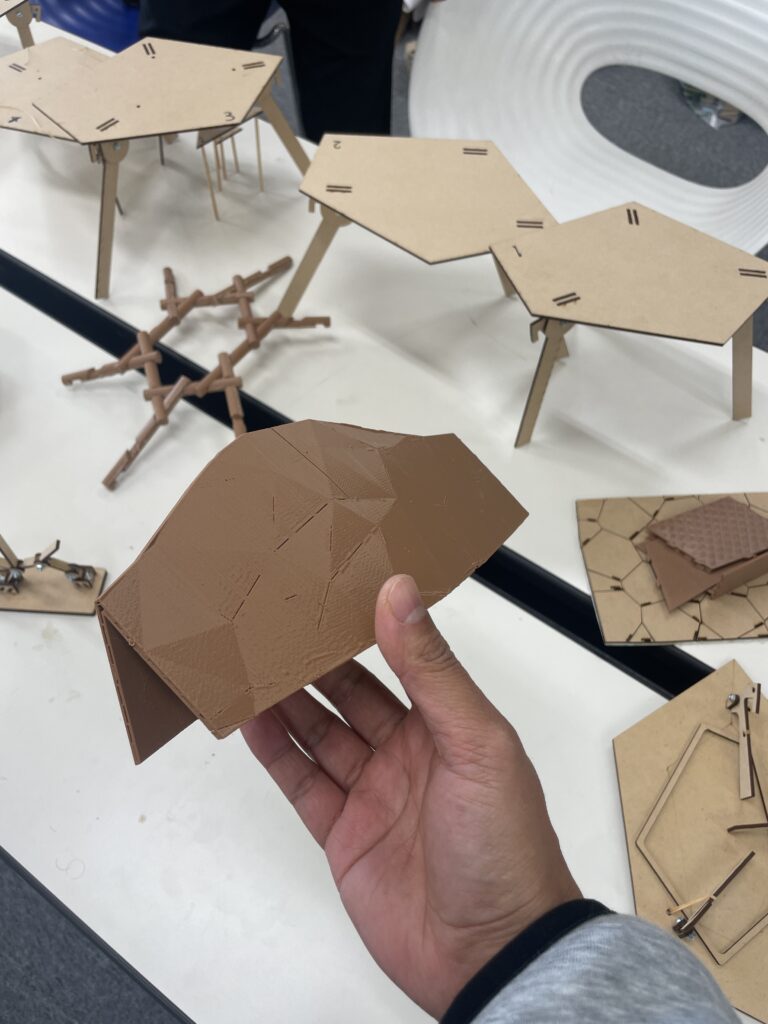 Close-up of a 3D-printed parametric structure held in hand, with cardboard architectural models on the table, part of a student project using Rhino3D and Grasshopper