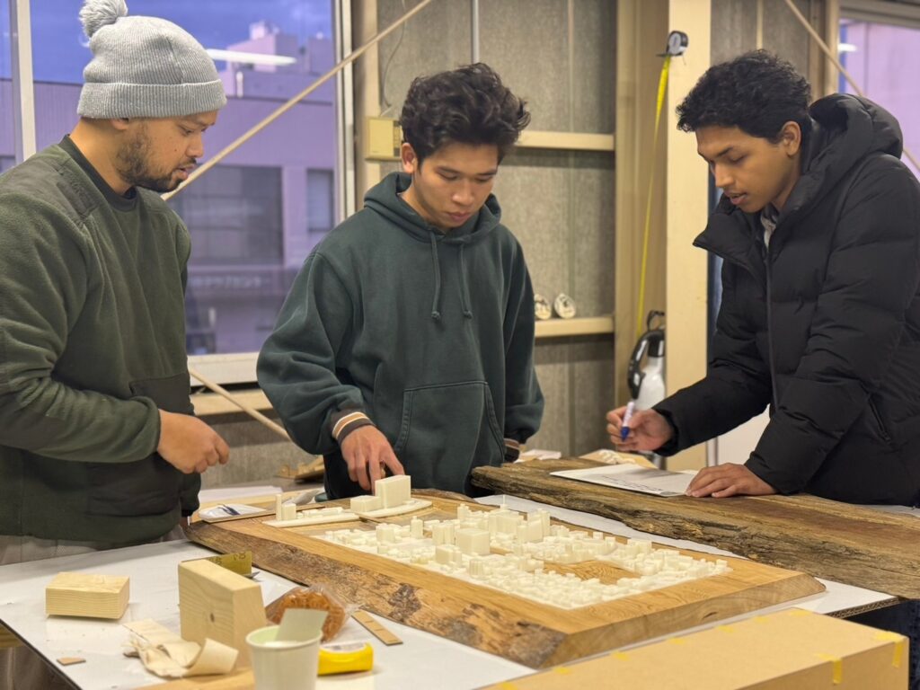 Architectural students collaborating on a site model with 3D-printed components mounted on a wooden base in a workshop setting